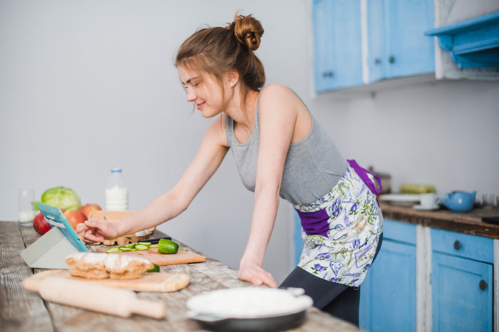 Young woman in apron cooking in kitchen, using a tablet with mom-boyfriend-food-drama theme in focus Young woman in apron cooking in kitchen, using a tablet with mom-boyfriend-food-drama theme in focus