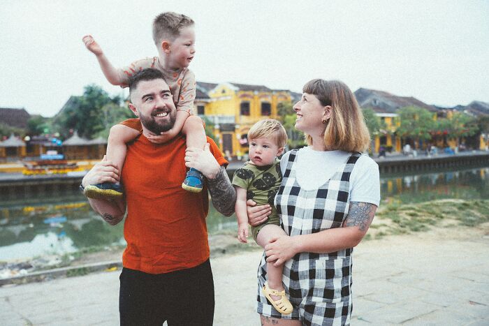 Family outdoors by the water with mom looking upset and husband smiling while carrying children on a sunny day