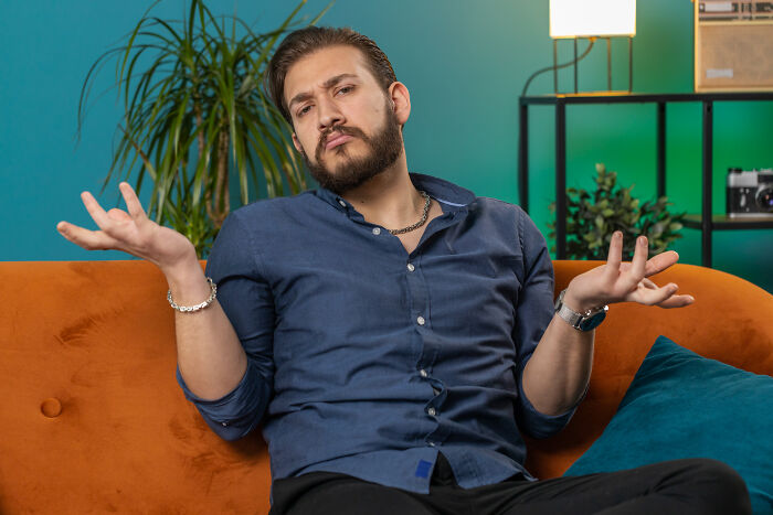 Man sitting on an orange couch with arms raised, showing a frustrated expression about secret funds in a home setting. Man sitting on an orange couch with arms raised, showing a frustrated expression about secret funds in a home setting.
