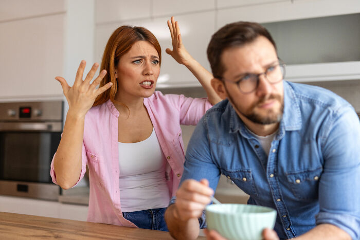Upset mom gesturing angrily at husband in kitchen, who looks away while holding a bowl, hinting at secret funds conflict. Upset mom gesturing angrily at husband in kitchen, who looks away while holding a bowl, hinting at secret funds conflict.