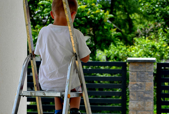 Young child climbing a ladder in a family backyard with greenery and a stone pillar fence in the background.