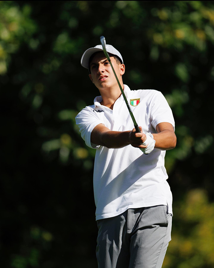 Young golfer in white shirt and cap swinging a club on a sunny day with blurred green trees in the background.