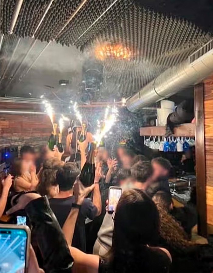 Crowded Swiss ski bar with people holding champagne bottles and sparklers, possibly linked to negligence causing lethal fire. Crowded Swiss ski bar with people holding champagne bottles and sparklers, possibly linked to negligence causing lethal fire.
