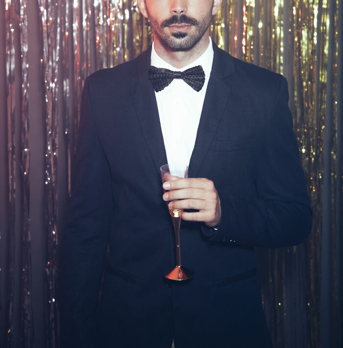 Man in a suit and bow tie holding a champagne glass at a nightclub party with shiny gold and silver backdrop.