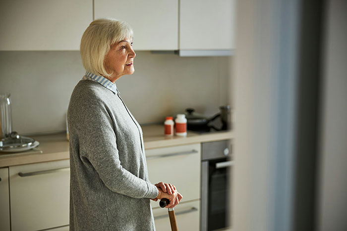 Older woman standing in kitchen with a cane, reflecting on family and retirement plan expectations. Older woman standing in kitchen with a cane, reflecting on family and retirement plan expectations.