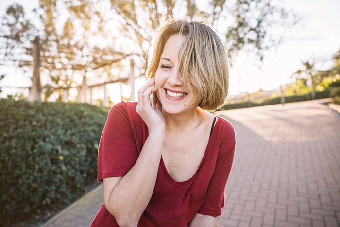 Young woman in a red shirt smiling and talking on the phone outdoors, representing niece in retirement plan story. Young woman in a red shirt smiling and talking on the phone outdoors, representing niece in retirement plan story.