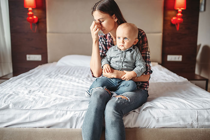 Woman sitting on bed holding toddler son, looking stressed, illustrating conflict over paternity test and family therapy suggestion. Woman sitting on bed holding toddler son, looking stressed, illustrating conflict over paternity test and family therapy suggestion.