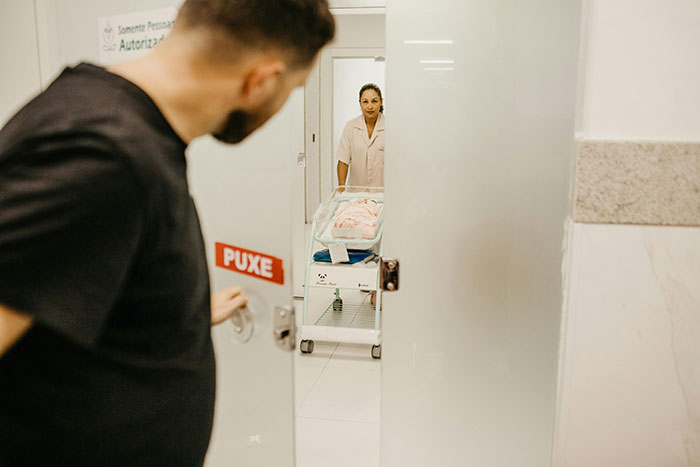 Man opening hospital door looking at newborn baby in a bassinet with nurse nearby, a scene of newborns not resembling the father.