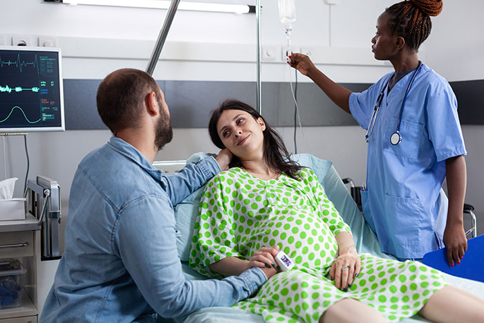Pregnant woman in hospital bed holding partner's hand while nurse adjusts IV, illustrating newborns who look nothing like father.