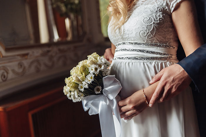 Pregnant woman in a white lace dress holding flowers, with a man's hands gently resting on her belly.