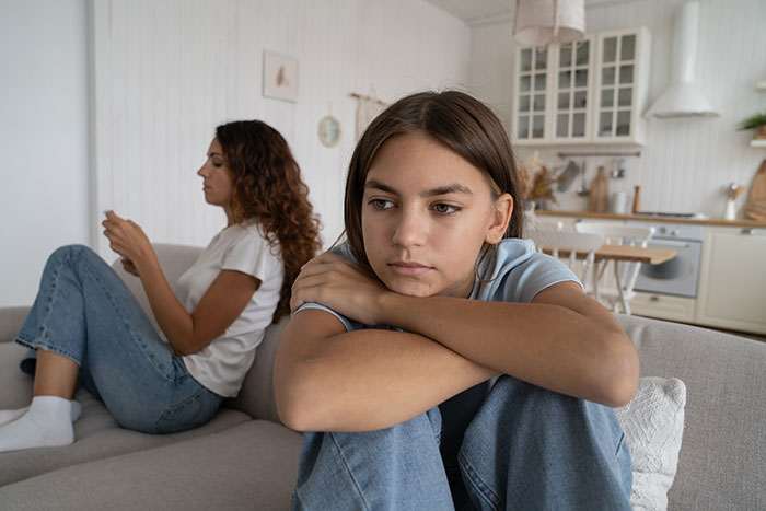 Two women sitting apart on a couch, one looking upset while the other uses her phone, illustrating relationship tension. Two women sitting apart on a couch, one looking upset while the other uses her phone, illustrating relationship tension.