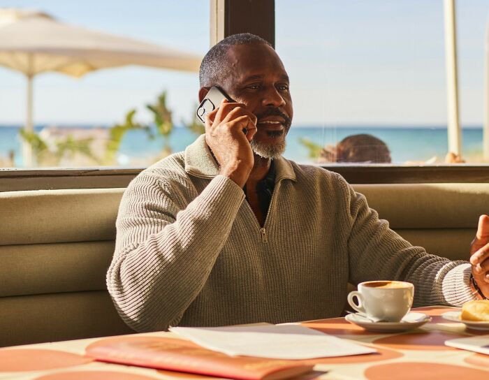 Man in a beige sweater talking on phone at a cafe table with coffee and pastry, capturing awkward moments in the service industry.