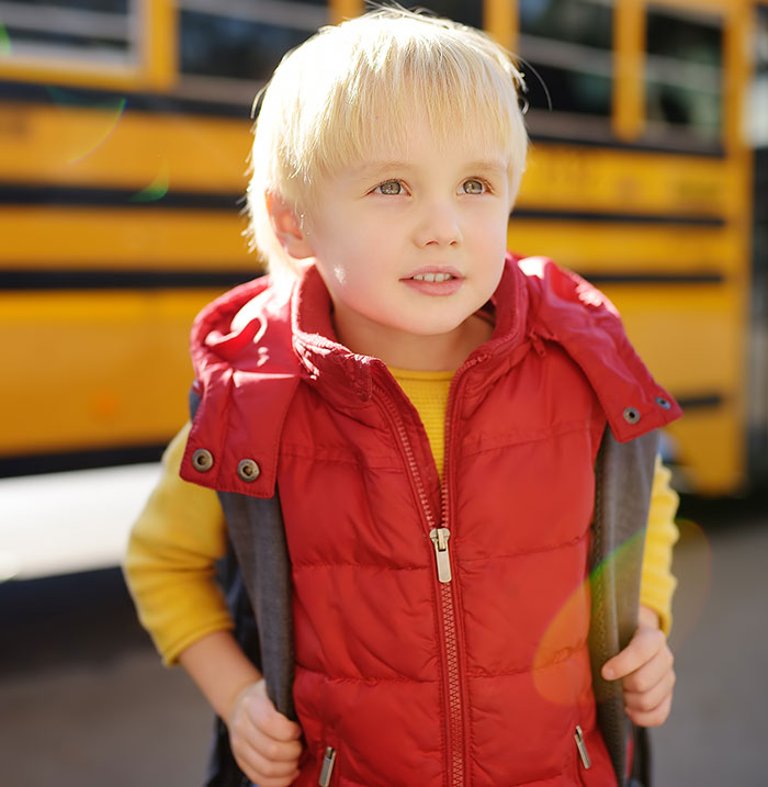 Young child wearing a red vest and yellow shirt standing near a yellow school bus, illustrating parenting differences around the world.