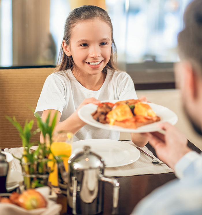 Young girl smiling and sharing food with an adult at breakfast, illustrating parenting differences around the world.
