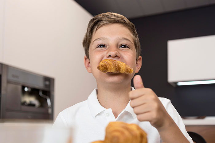 Young boy holding a croissant in his mouth and giving a thumbs up indoors showing parenting differences around the world.