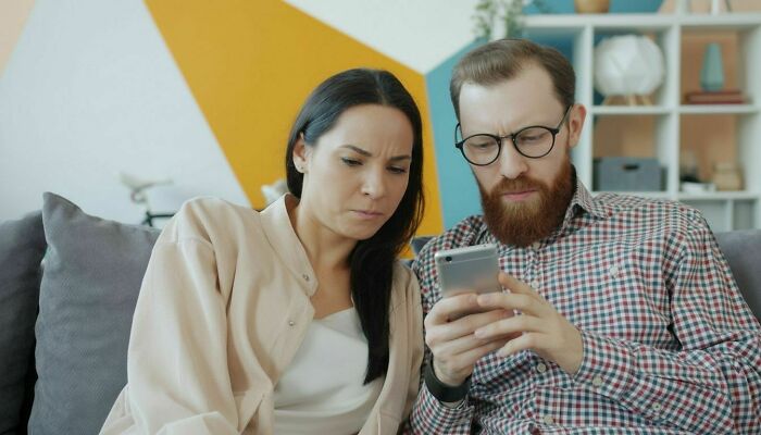 Woman furious and partner looking at phone on couch, upset about family laughing at their toddler Woman furious and partner looking at phone on couch, upset about family laughing at their toddler