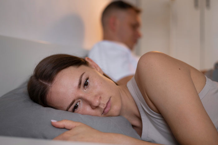 Woman lying awake in bed looking worried while her partner sleeps in the background, partner sleep called ambulance context.