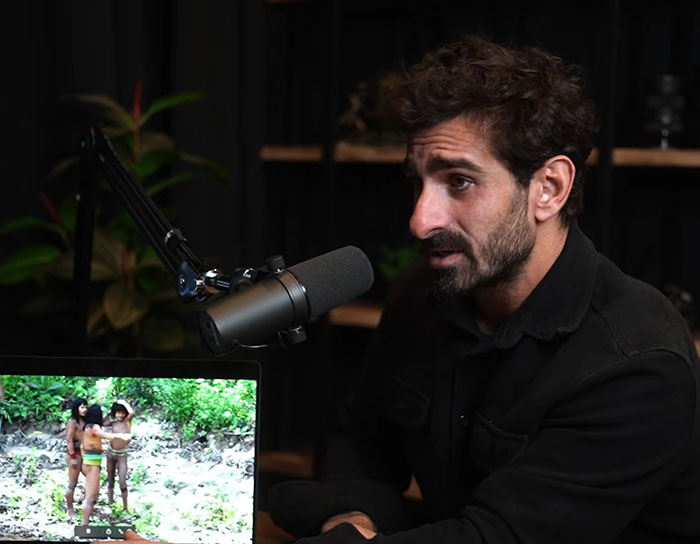 Man speaking into a microphone in a dark room with footage of an uncontacted Amazonian tribe on a laptop screen.