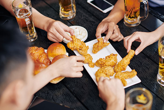 People grabbing fried chicken from a platter at a restaurant, enjoying a casual meal with drinks and bread rolls.