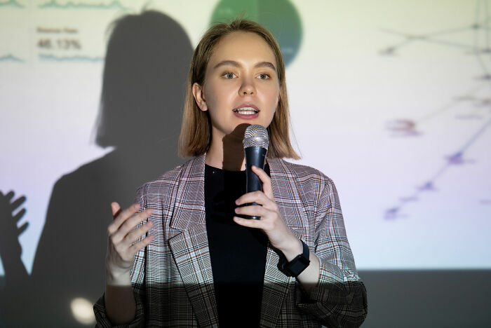 Young woman presenting practical life hack cheat codes using a microphone against a business data presentation background.