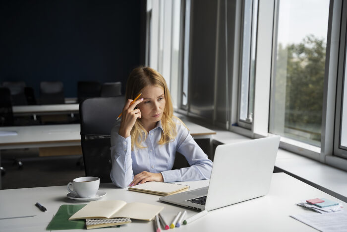 Woman working on laptop in office, thinking deeply while holding pencil, surrounded by notebooks and a coffee cup, real-life cheat codes.