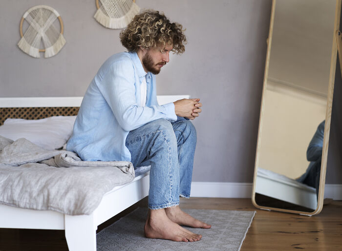 Man sitting on bed, barefoot and thoughtful, reflecting in a mirror, representing real-life cheat codes concept.