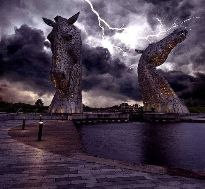 Two large horse head sculptures by water under a stormy sky with lightning, a powerful photo from a country.