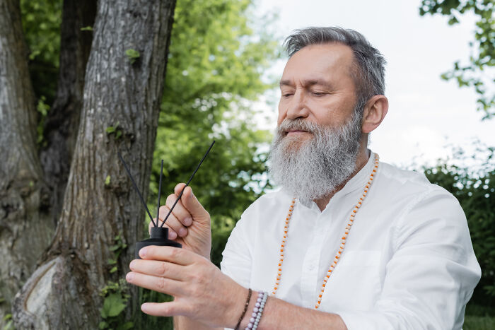 Bearded man in white shirt holding incense sticks outdoors, reflecting on stories about the worst person they've met.