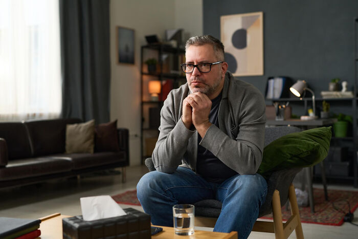 Middle-aged man in glasses sitting thoughtfully in living room, reflecting on strange tales of people who morphed unexpectedly.