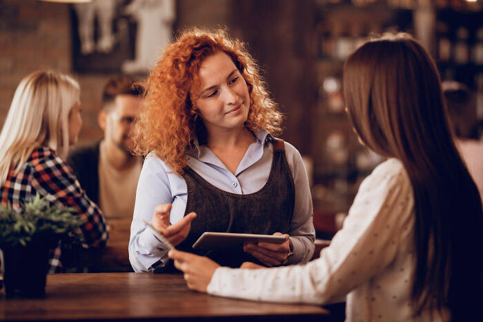 A woman with red curly hair talking to another woman at a table, illustrating strange tales of folks who morphed.