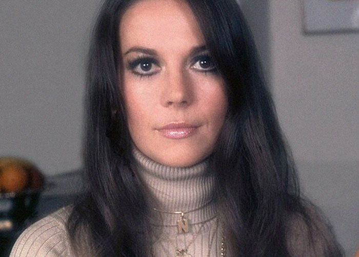 Close-up of a woman with long dark hair wearing a beige turtleneck and necklace, related to celebrity passings.