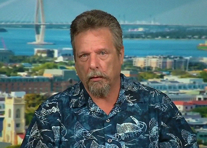 Man with goatee wearing a blue patterned shirt speaking on camera with cityscape and bridge in the background about celebrity passings.