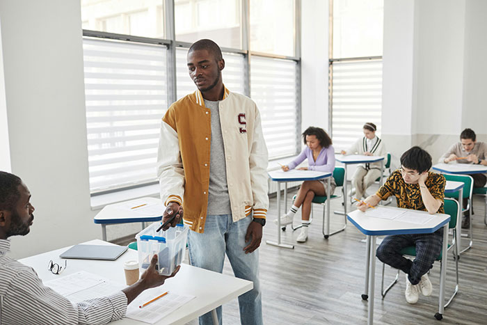 Student handing phone to professor enforcing phone rule in classroom while classmates focus on exam or assignments.
