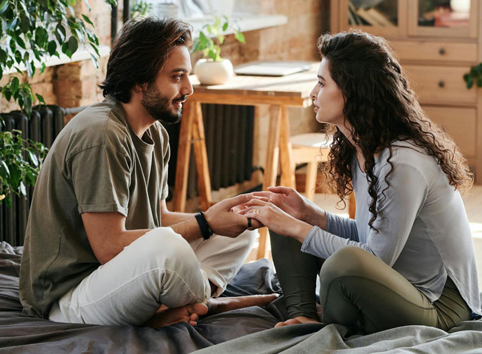Man and woman sitting on bed holding hands, discussing relationship concerns about having kids and honesty. Man and woman sitting on bed holding hands, discussing relationship concerns about having kids and honesty.