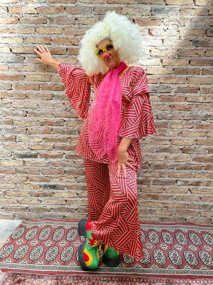 Clown with white wig, yellow glasses and oversized shoes posing by brick wall, rained frogs.