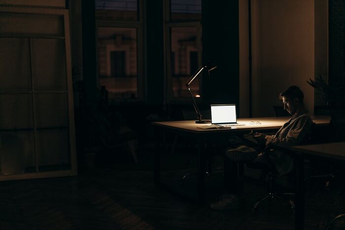 Worker on a night shift sitting near a desk with a laptop and lamp, reflecting 3 AM chaos during overnight work hours.