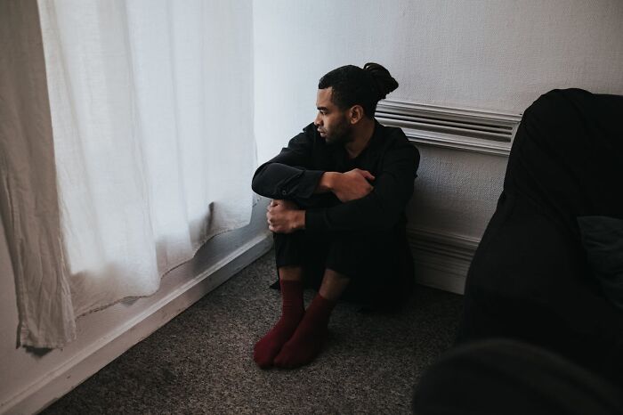 Man sitting on carpeted floor by a window, hugging knees and staring out, wearing black clothes and red socks, rained frogs.