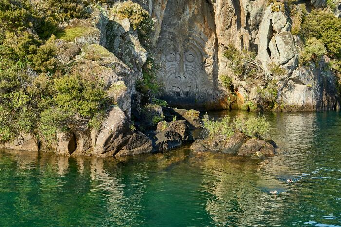 Rock carving of a face on a cliff beside a green lake with swimmers, showcasing lesser-known facts about countries.