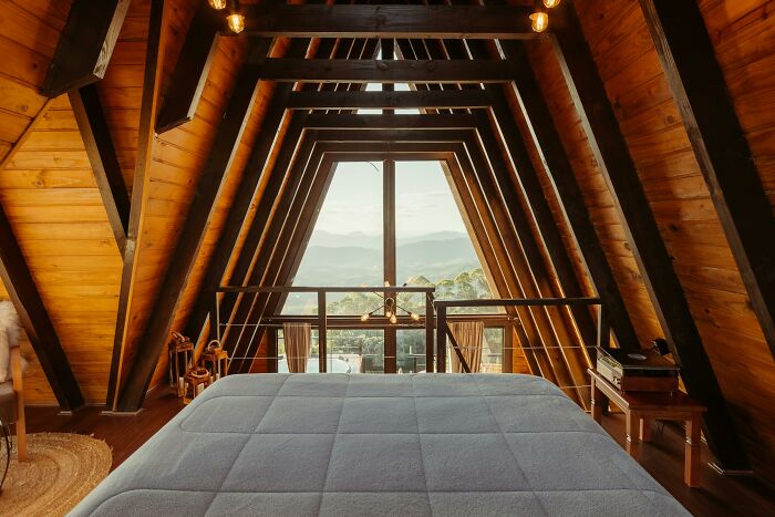 Cozy wooden A-frame cabin bedroom with a large window showcasing mountain views and warm natural lighting.