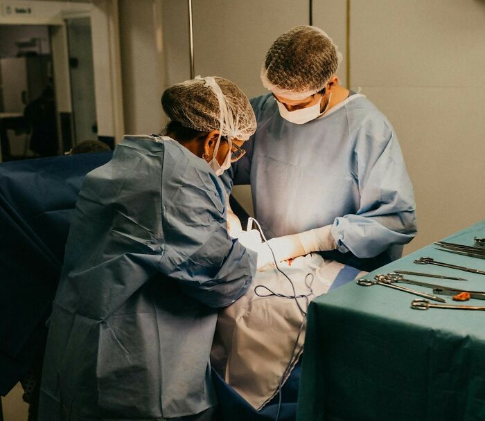 Two hospital workers in surgical gowns and masks performing an operation under bright light, instruments on table.