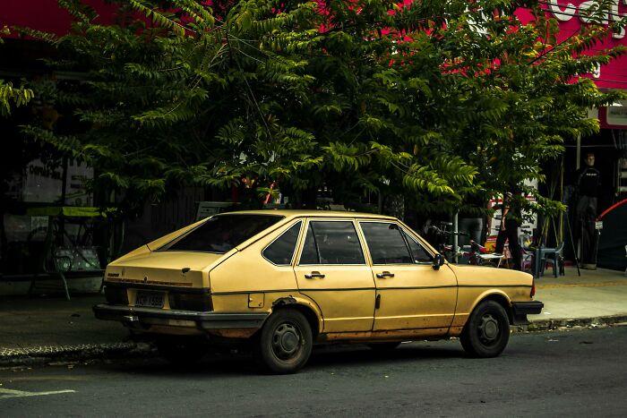 Old yellow car parked on a city street with trees overhead, illustrating contrast to rich people’s out of pocket statements.