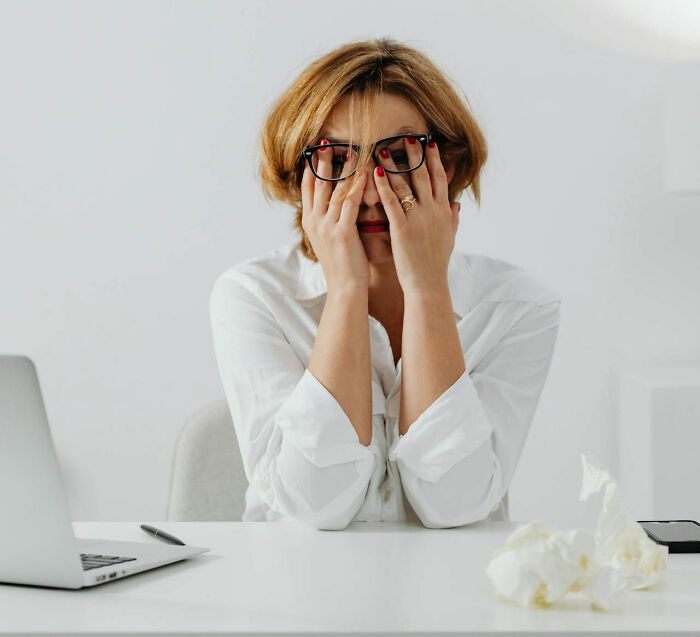 Stressed woman in glasses at desk with laptop and tissues, covering face as if it had rained frogs