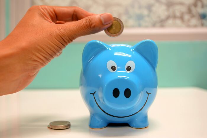 Hand placing a coin into a blue piggy bank representing saving money and wealth from rich people’s out of pocket comments.