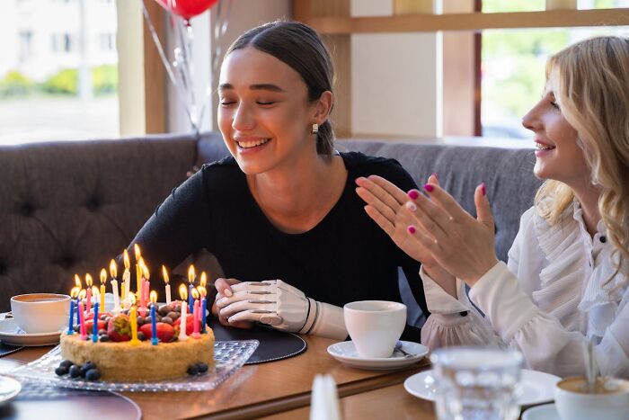 Young woman with a prosthetic hand smiling by a birthday cake while a friend claps, capturing awkward moments service industry workers face.