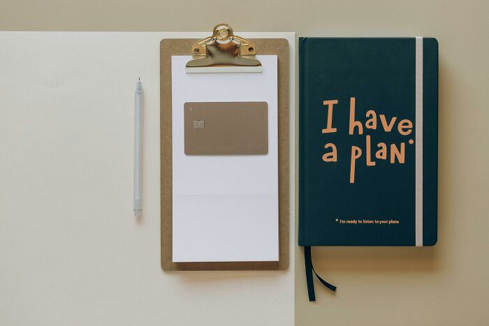 Clipboard with credit card, pen, and dark green planner with I have a plan text on beige background representing wealth and finances.