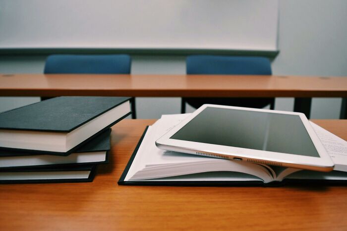 Tablet resting on an open book with stacked notebooks on a wooden desk in an empty classroom setting.