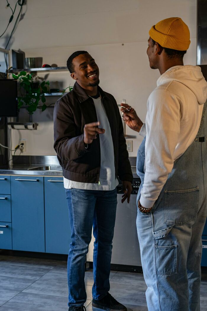 Two men laughing and talking in a kitchen, one wearing a yellow beanie and overalls, rained frogs