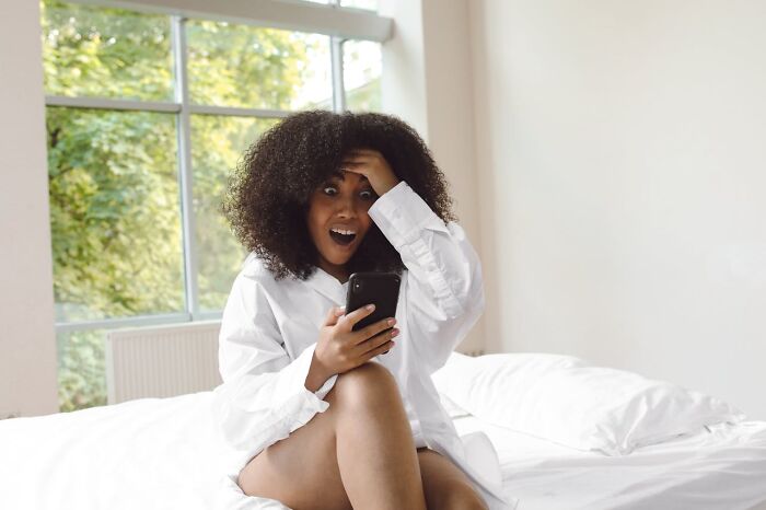 Woman with curly hair in white shirt sitting on bed, looking shocked at phone screen after accidental text causing panic.