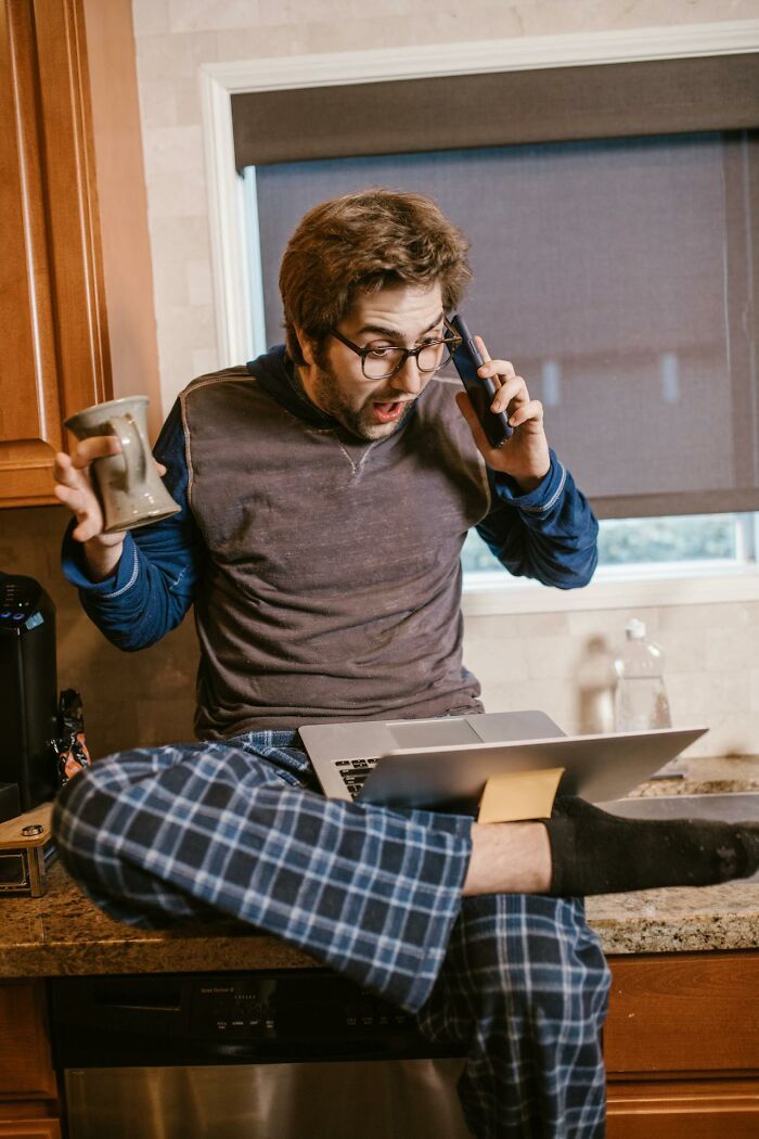 Man in pajamas with glasses looking shocked on phone call while using laptop, illustrating accidental texts panic reaction.