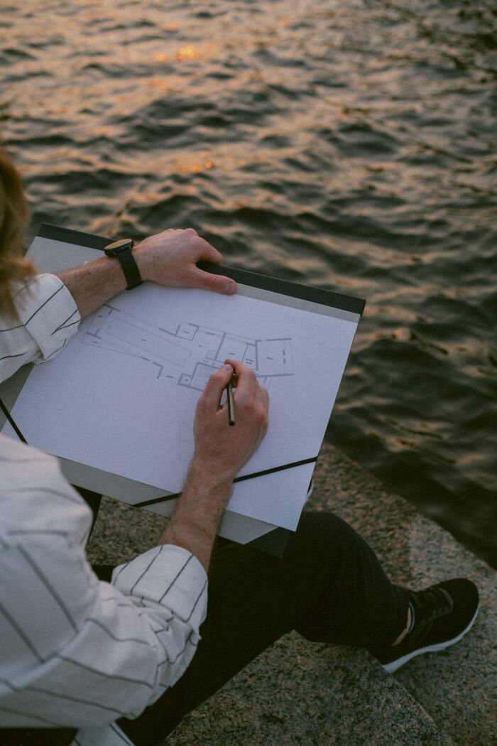 Person sketching building plans by the water at sunset, suggesting creepiest displays of intelligence.